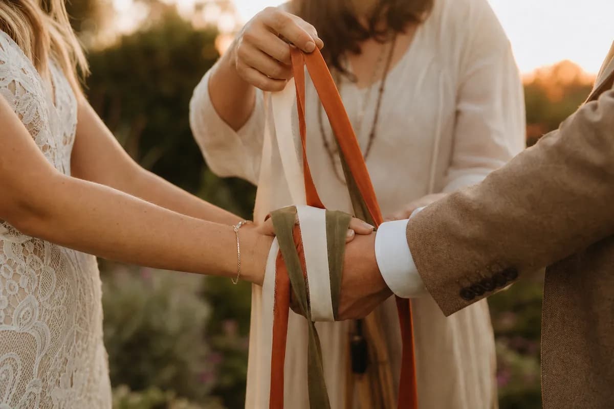 Manos entrelazadas con listones terracota, crema y olivo en ritual de handfasting al atardecer