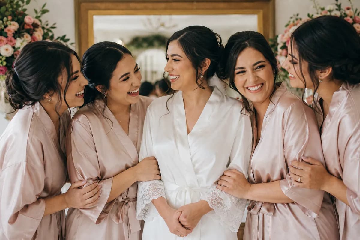Novia y damas de honor en batas a juego riendo frente al espejo mientras se arreglan por la manana