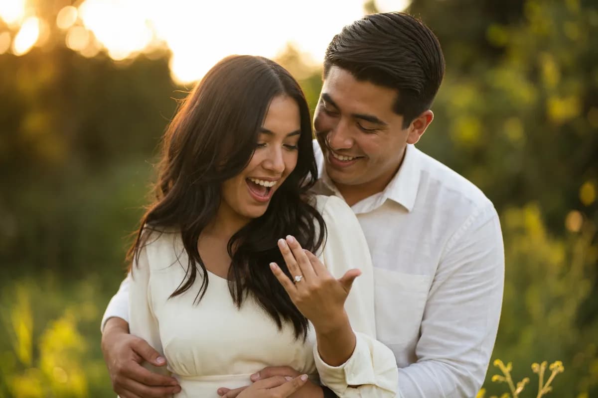 Pareja mexicana riendo en jardín al atardecer; ella mira feliz su anillo de compromiso recién puesto
